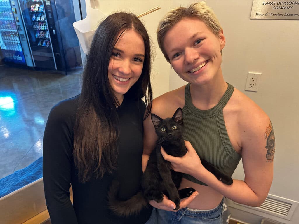 Two women stand indoors holding a small black kitten, with a doorway and wall sign in the background.