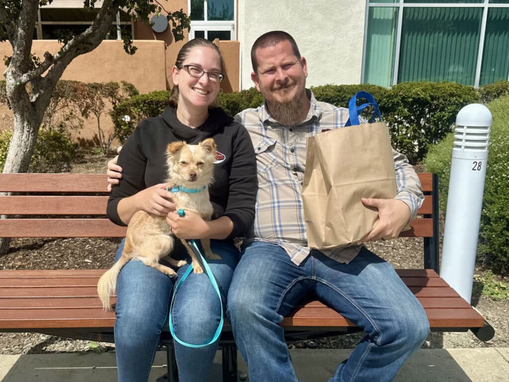 A woman and a man sit on a bench holding a small tan dog, with a brown paper bag beside them and a building with windows in the background.
