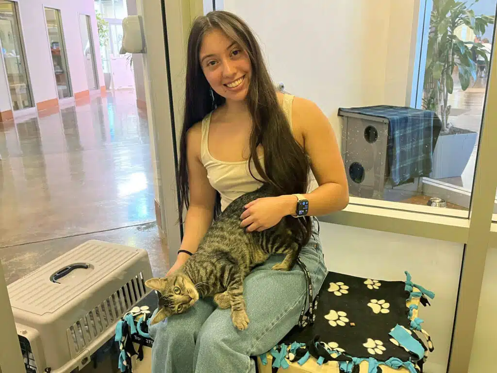 A smiling woman sits on a patterned blanket bench holding a tabby cat, with a pet carrier on the floor and a windowed indoor lobby behind her.