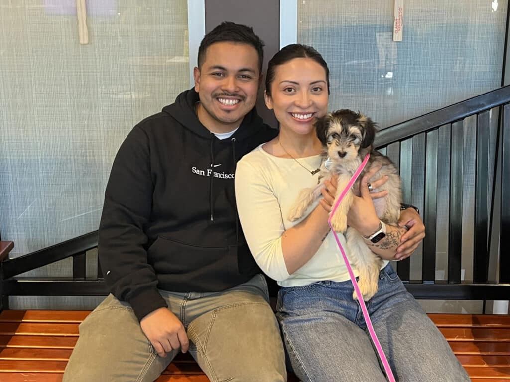 A smiling couple sits on a bench holding a small puppy with a pink leash, in front of a glass wall and black railing.