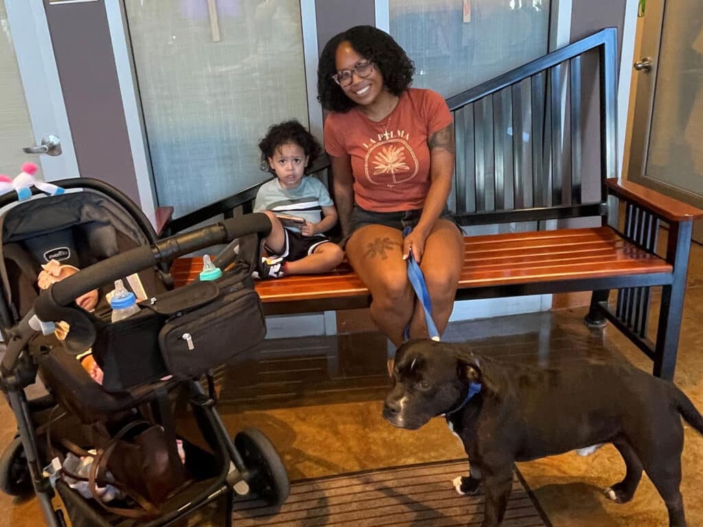 A woman sits on a wooden bench beside a toddler, with a black dog standing in front and a stroller in the foreground.
