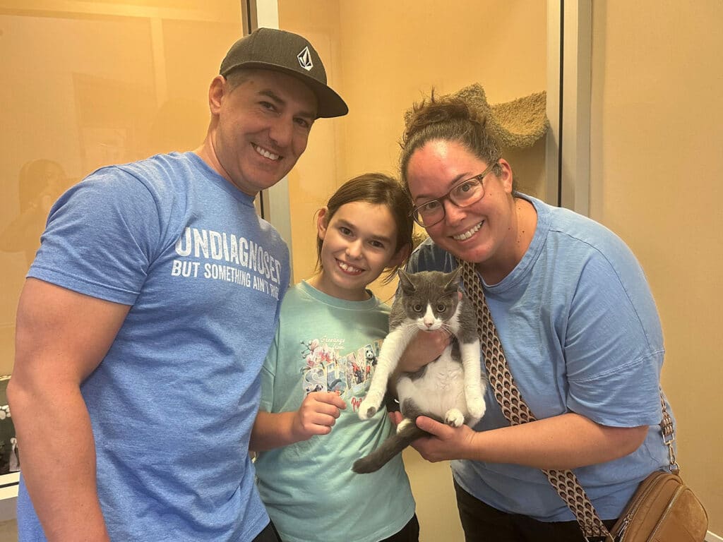 A man, a girl, and a woman hold a gray-and-white cat indoors, with beige walls and a doorway in the background.