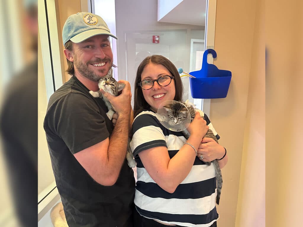 A smiling man in a baseball cap and a woman in glasses hold two cats indoors, with a blue wall-mounted accessory and beige walls in the background.