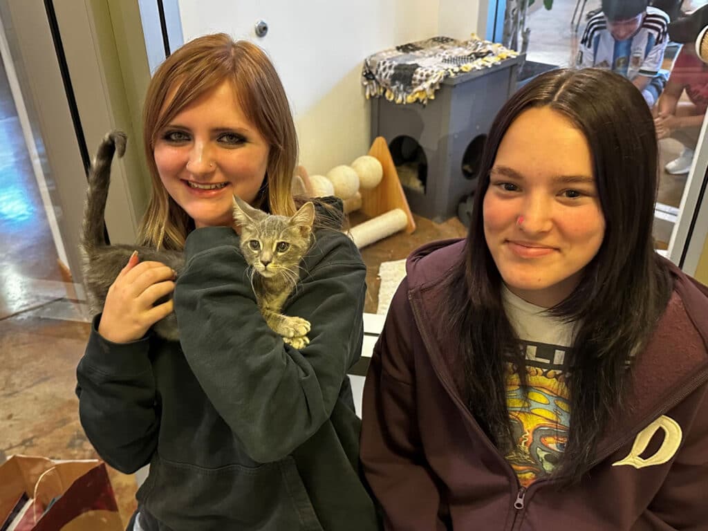 Two young women stand indoors holding a small tabby kitten, with a cat tree and pet supplies in the background.