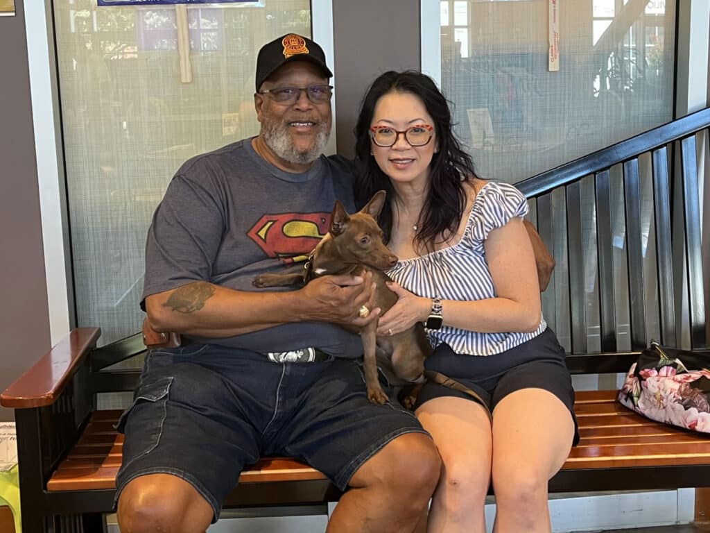 A man and woman sit on a wooden bench holding a small brown dog, with a windowed wall and staircase behind them.