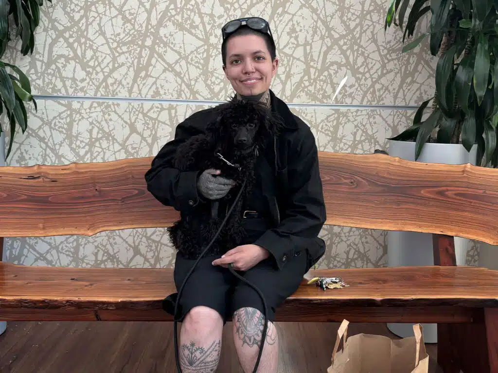A woman in a black outfit sits on a wooden bench holding a small black dog, with potted plants and patterned wallpaper behind her.