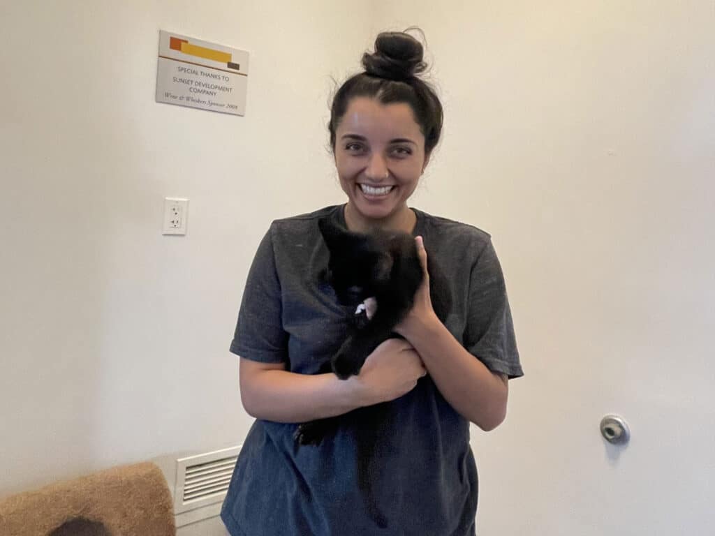 A woman in a gray T-shirt holds a small black kitten in front of a plain white wall, with a vent and wall sign in the background.