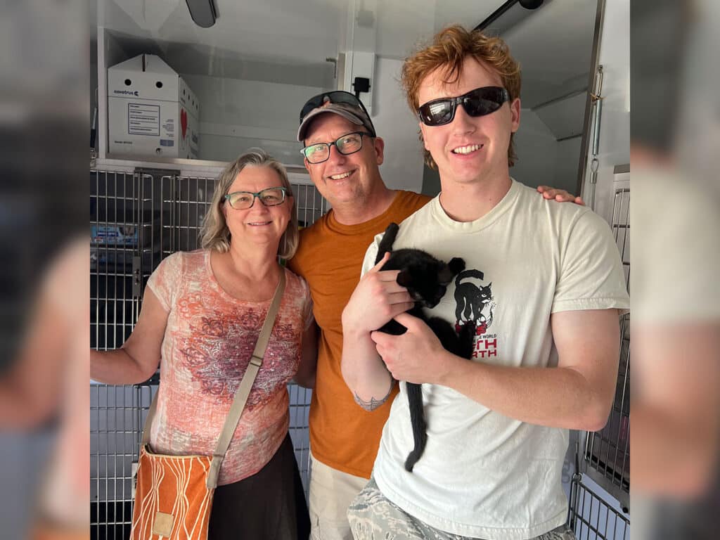 Three adults stand inside a pet transport crate area, with the person in front holding a small black kitten and metal cages visible behind them.