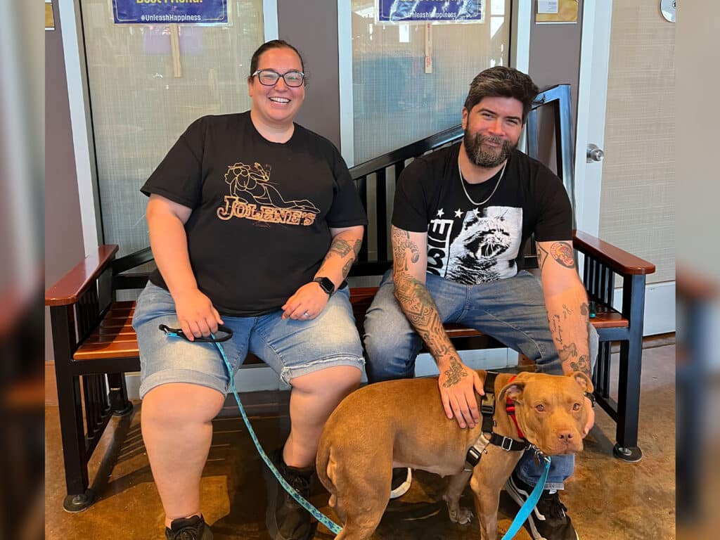Two people sit on a bench indoors with a brown dog on a leash in front of them, as the man pets the dog beside glass doors.