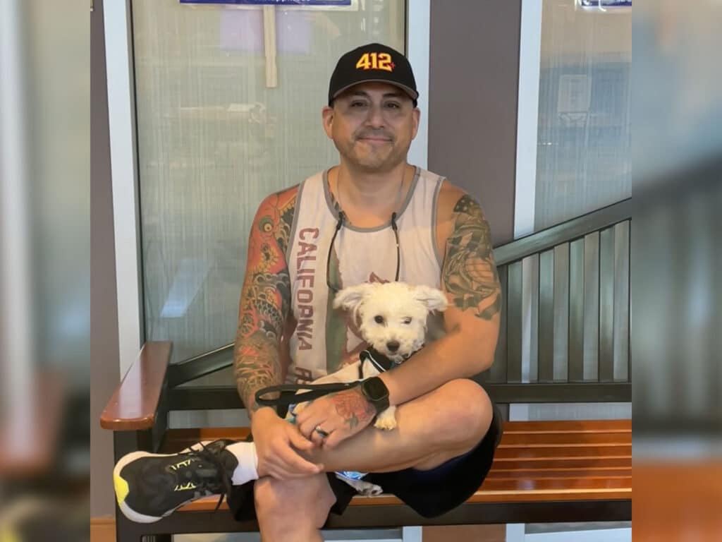 A tattooed man wearing a black cap and sleeveless shirt sits on a bench holding a small white dog, with glass walls and metal railings behind them.