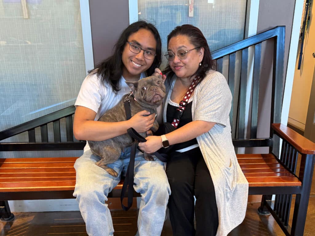 Two women sit on a wooden bench holding a gray French bulldog, with glass doors and a stair railing behind them.