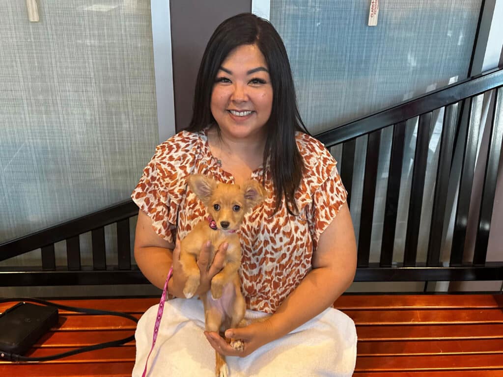 A woman in a patterned blouse holds a small tan puppy with a pink leash while sitting on a wooden bench, with a black railing and blue wall behind them.