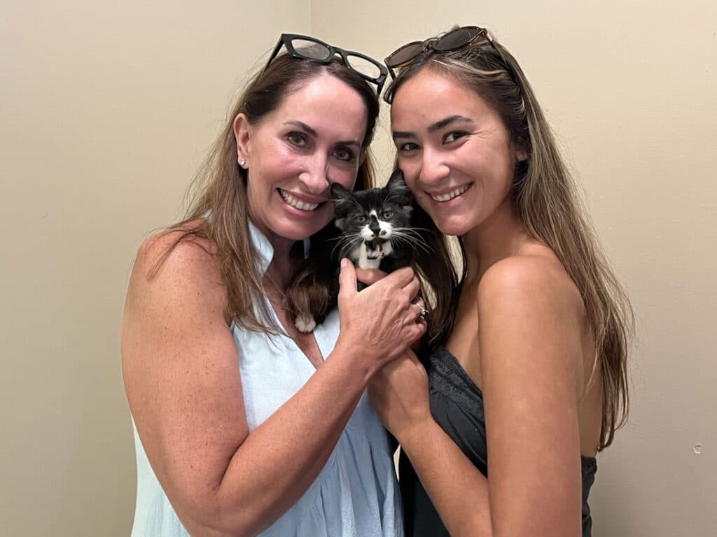 Two women hold a small black-and-white kitten in front of a beige wall.