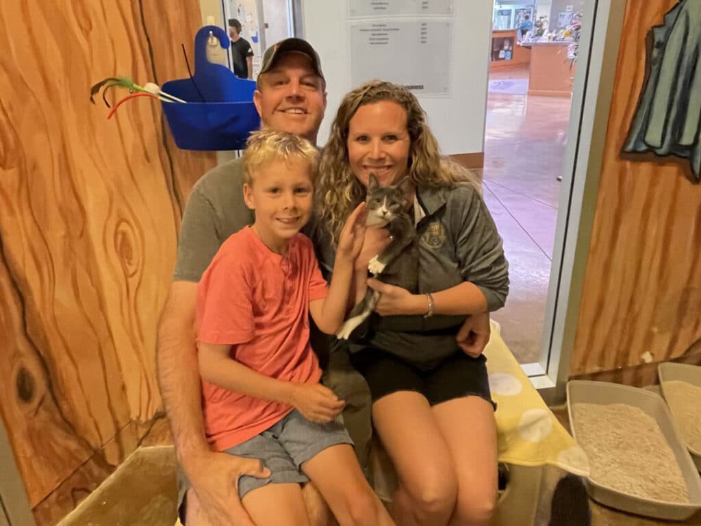 A family sits on a bench holding a gray-and-white kitten inside a pet adoption area with wood-paneled walls and a doorway in the background.