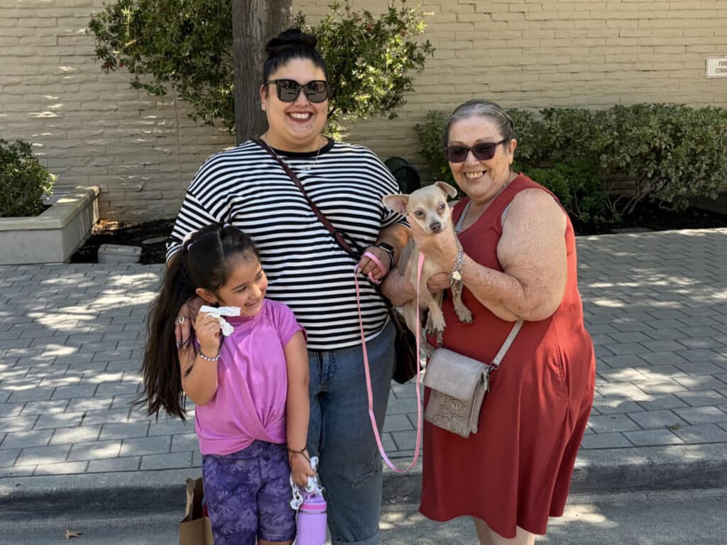 Three people stand outdoors on a paved walkway, with one woman holding a small tan dog on a pink leash.