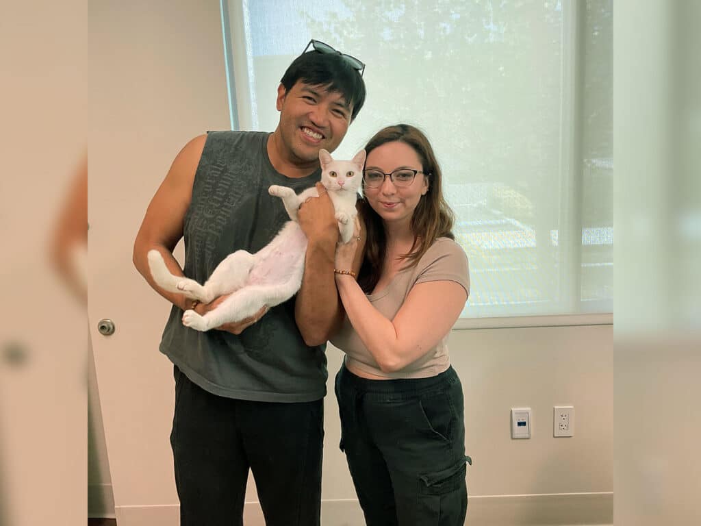 A man and woman hold a white cat in front of a large window in a bright room.