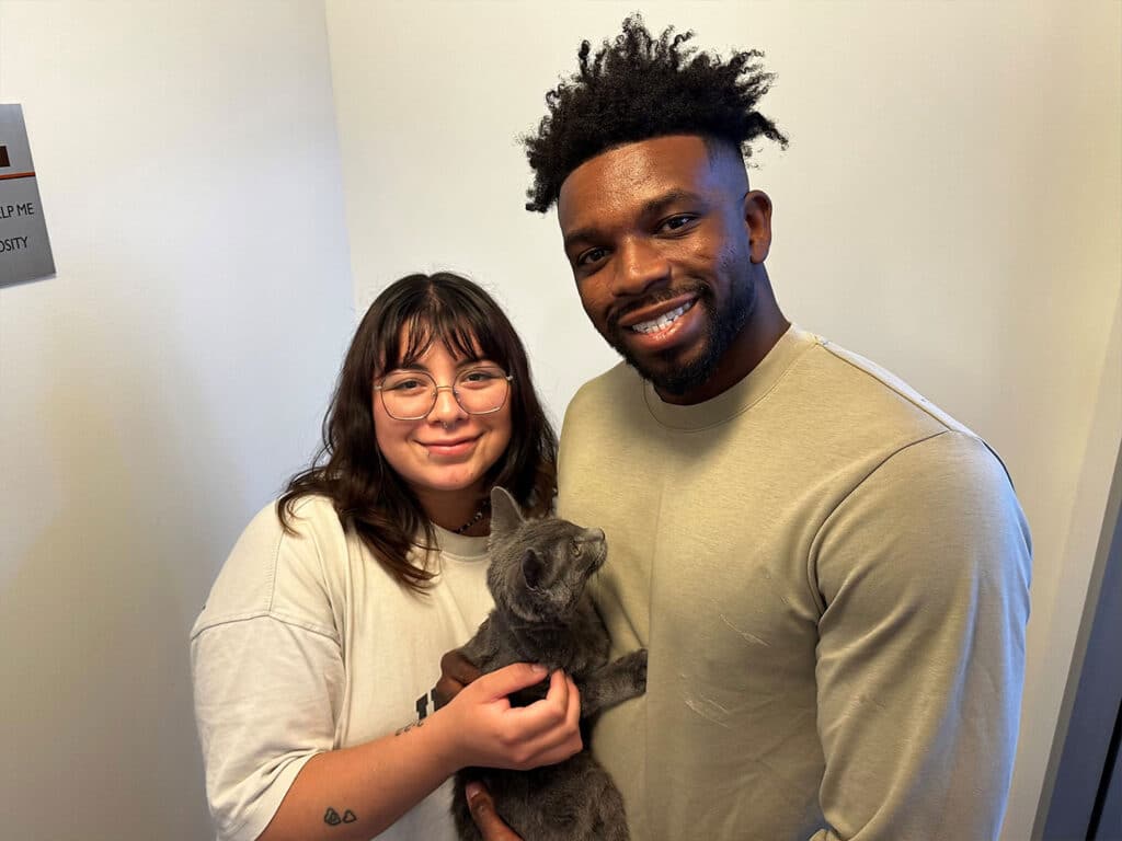 A smiling couple holds a gray cat indoors against a plain white wall, with a small sign partially visible on the left.