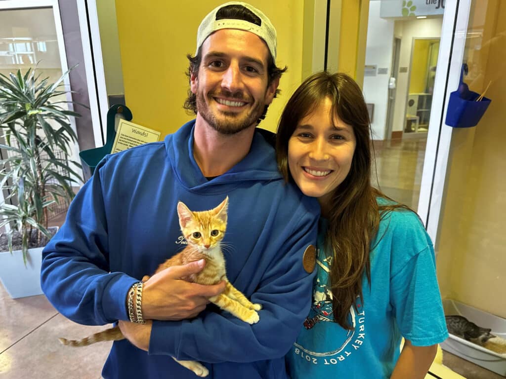 A smiling man in a blue hoodie and a woman in a teal shirt hold a small orange kitten in a hallway with glass doors and potted plants.