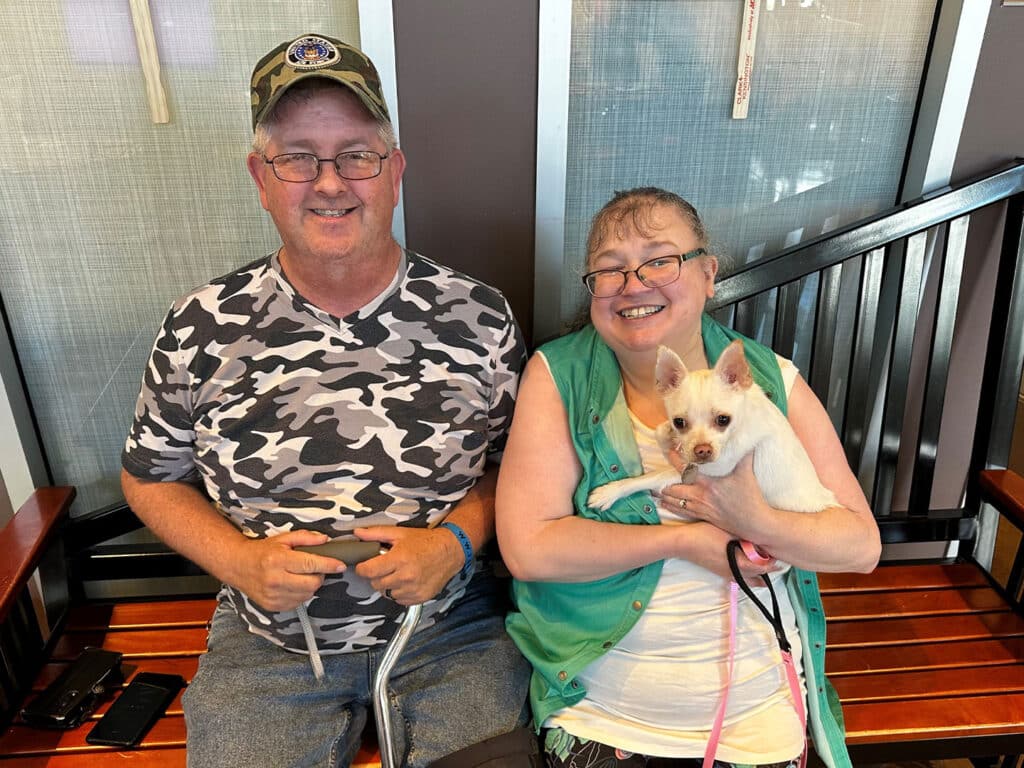 A smiling man in a camouflage shirt and cap sits beside a smiling woman holding a small white dog with a pink leash on a bench indoors.