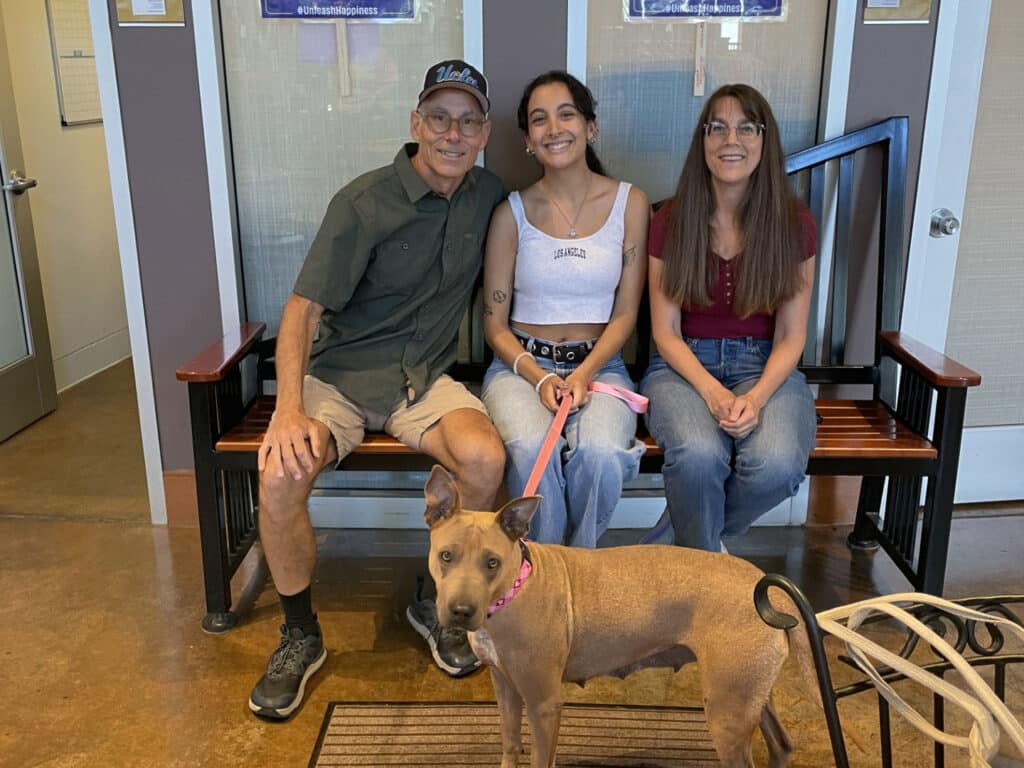 A tan dog wearing a pink collar stands in front of three people seated on a bench in an indoor lobby.