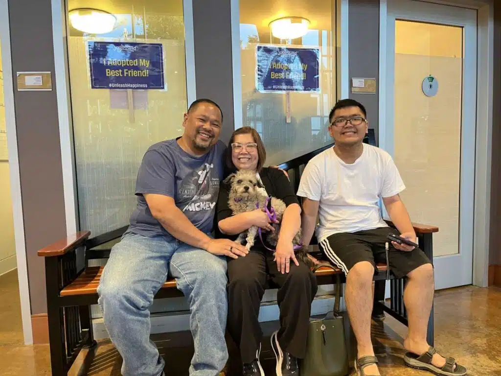 Three people sit on a bench with a small dog in the center, in front of glass doors with signs that read "Adopted My Best Friend."