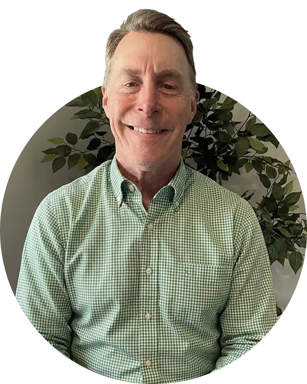 A smiling man in a green checkered button-down shirt poses in front of leafy plants, part of the Joybound Board of Directors.