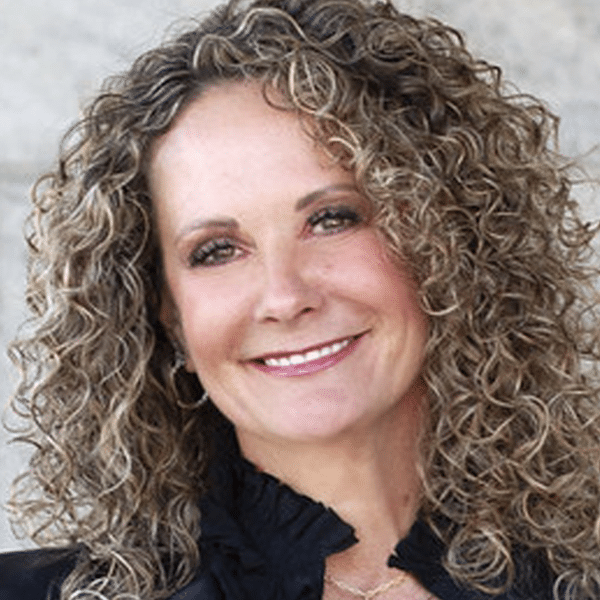 A smiling woman with curly light brown hair wears a black top against a plain light background.