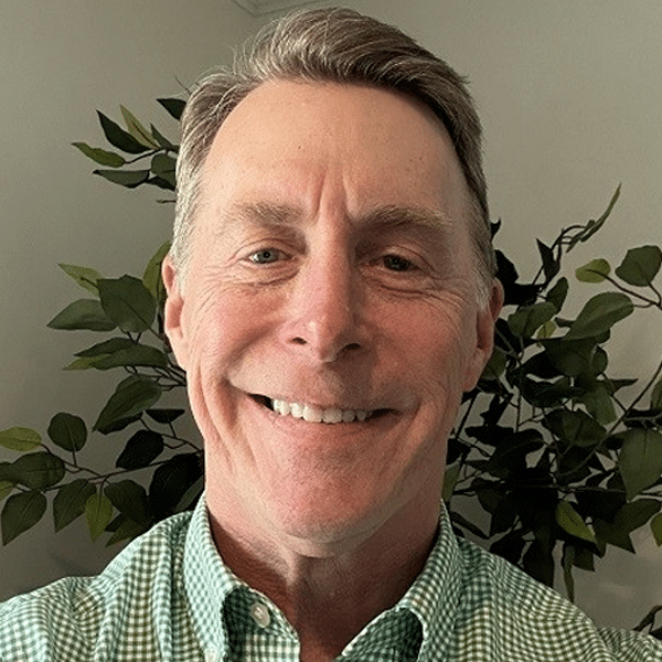 A smiling man in a green checkered shirt stands in front of leafy indoor plants against a plain wall.