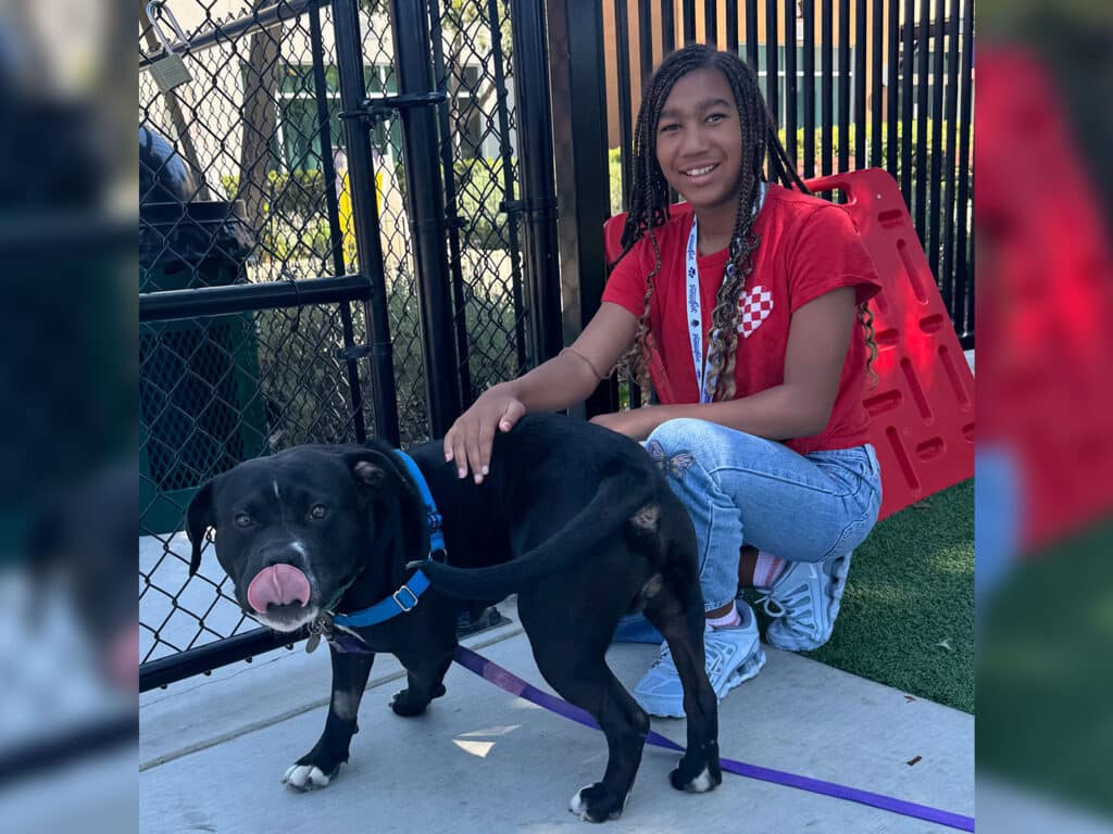 A smiling young woman in a red shirt kneels beside a black dog on a leash, with a chain-link fence and red barrier behind them.