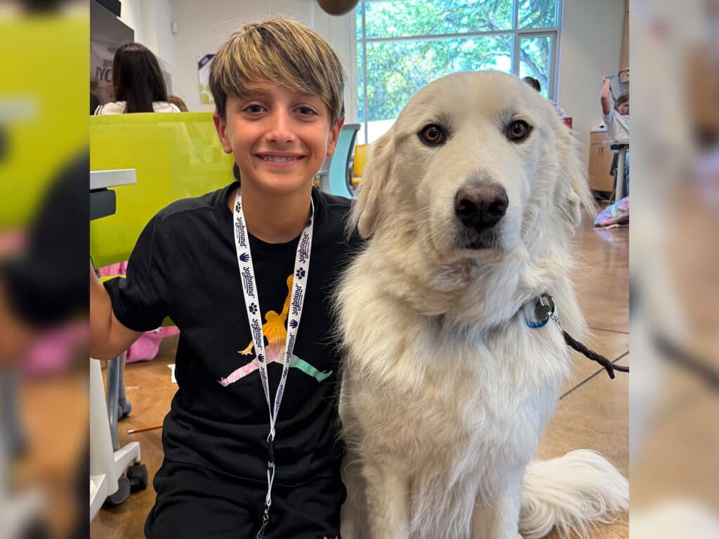 A smiling child with short blond hair sits beside a large white dog indoors, with a bright window and colorful partition in the background.