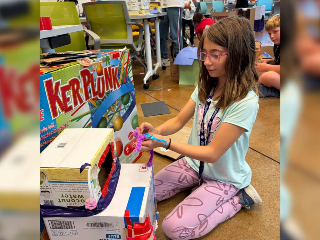A girl wearing glasses kneels on the floor and plays a KerPlunk game beside a cardboard craft setup in a classroom.