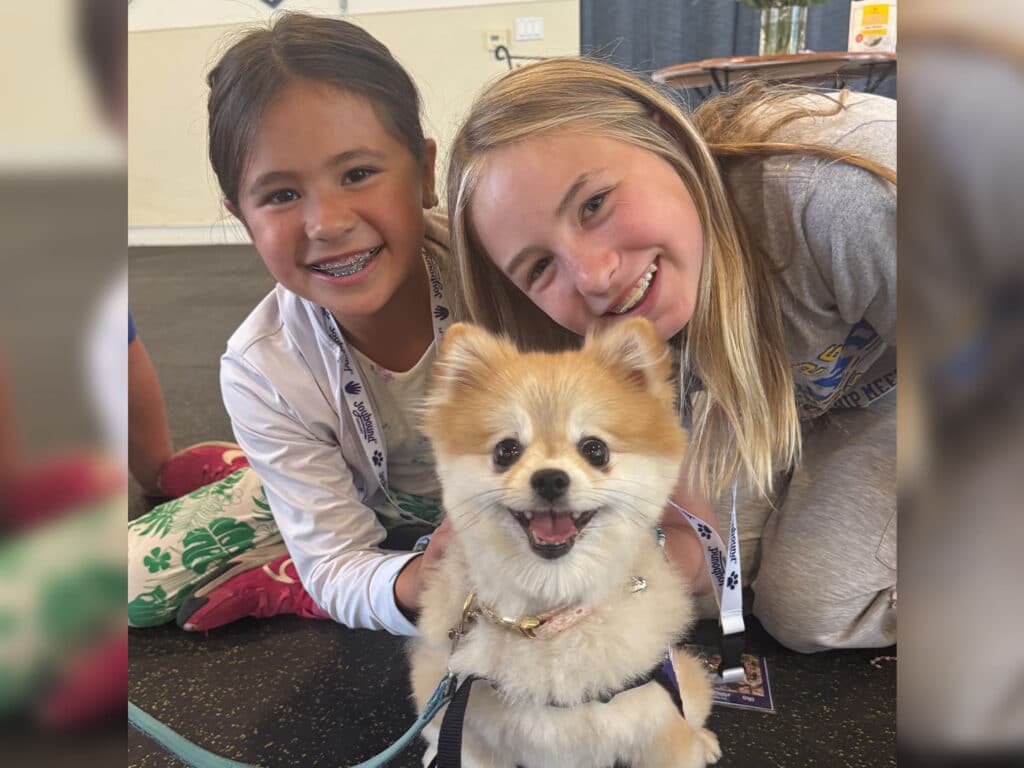 Two girls lean beside a small fluffy dog wearing a harness, with a camp room and scattered items in the background.