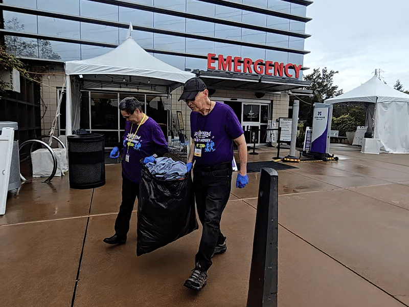 An older man and woman in purple shirts and blue gloves carry a black trash bag outside an emergency entrance, with white tents and a glass building in the background.