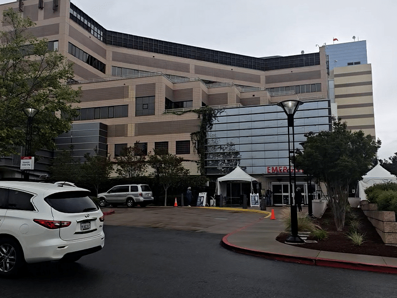 A large tan multi-story hospital building with glass windows, a curved driveway, parked cars, and a white entrance tent near the emergency department.