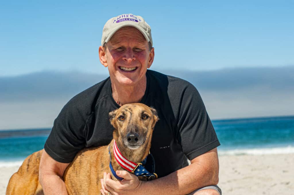 A man in a beige cap and black T-shirt holds a tan dog wearing a red, white, and blue collar on a sunny beach, with blue ocean water and sand behind them.