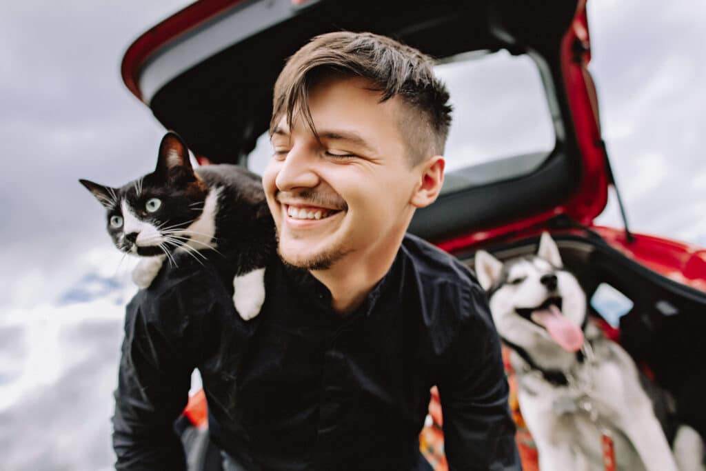 A smiling man holds a black-and-white cat on his shoulder beside an open red car trunk, with a husky in the background from the Joybound Strategic Plan.