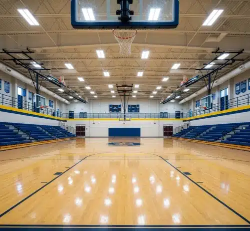 An empty indoor basketball gym with polished wood flooring, blue bleachers, and overhead lights beneath the ceiling.