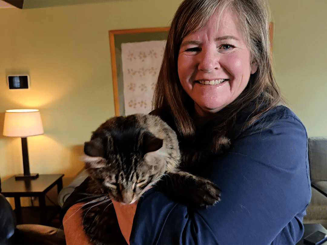 A smiling woman in a dark blue top holds a tabby cat indoors, with a lit table lamp and framed wall art in the background.