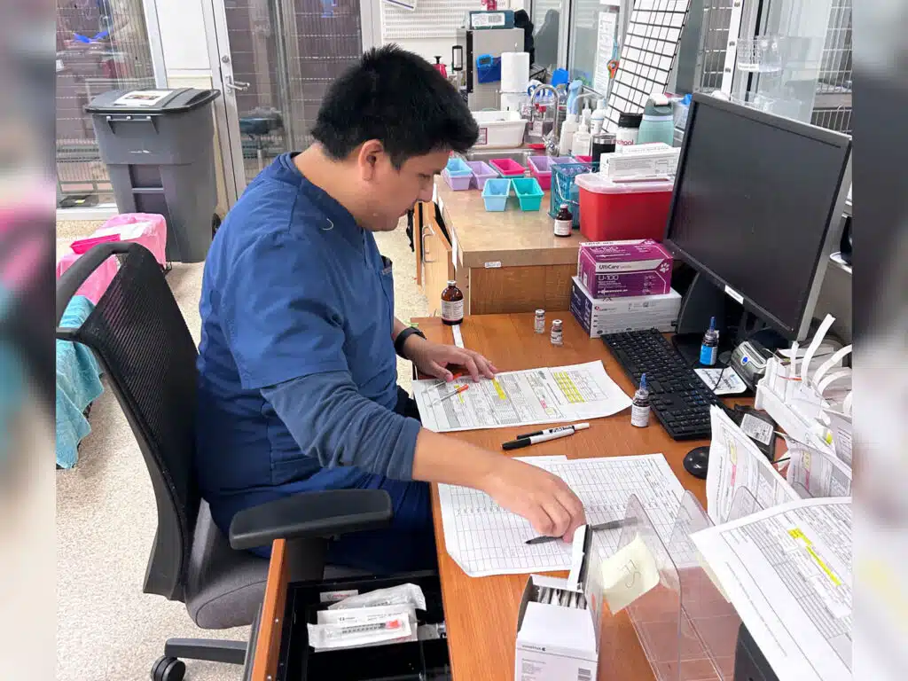 A man in blue scrubs reviews paperwork at a cluttered office desk with a computer monitor, keyboard, and supplies.