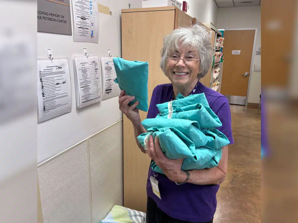 An older woman in a purple T-shirt holds several folded teal fabric bags in a hallway with posted notices and wooden doors.