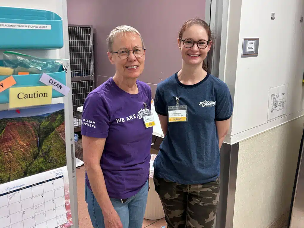 Two women wearing name badges stand indoors beside a wall and storage cabinet, with a caution sign and calendar visible on the left.