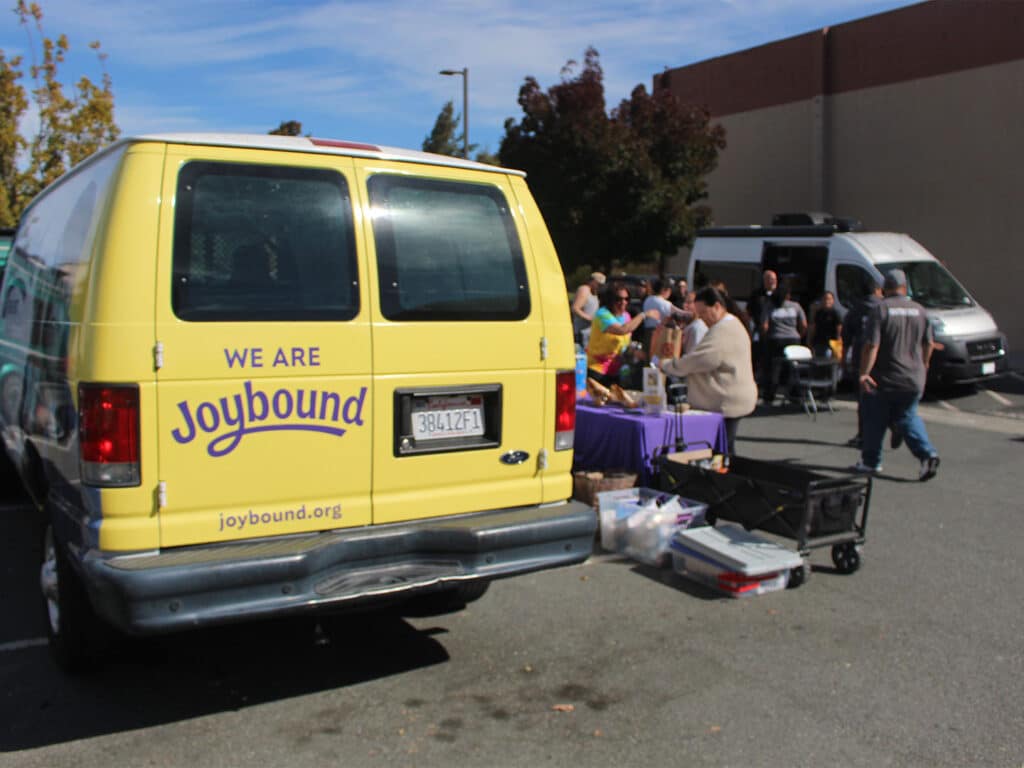 A yellow Joybound van is parked beside a food distribution table with volunteers and supplies, with a white van and shopping cart in the background.
