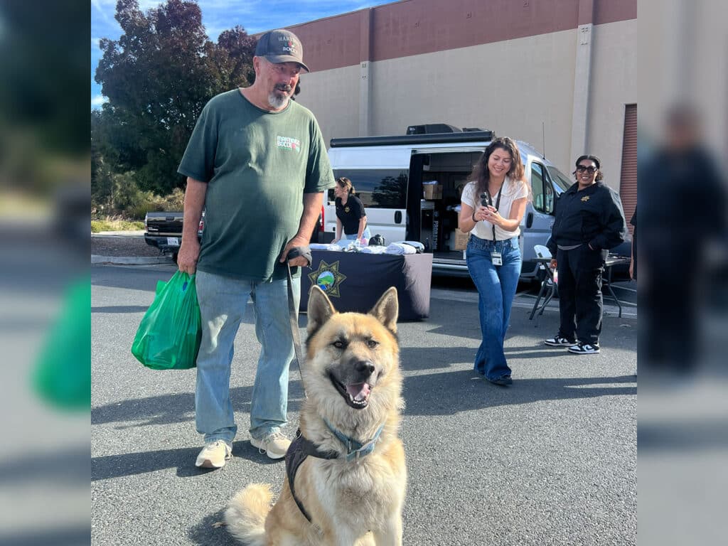 A man holding a green bag walks beside a tan dog in a harness, with volunteers and an open van at an outdoor FoodShare Pantry event.