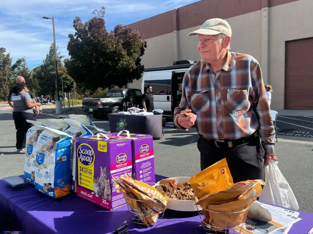 An older man in a plaid shirt and cap stands beside a purple-covered table with assorted food donations and bags, with people and vehicles outside a building in the background.