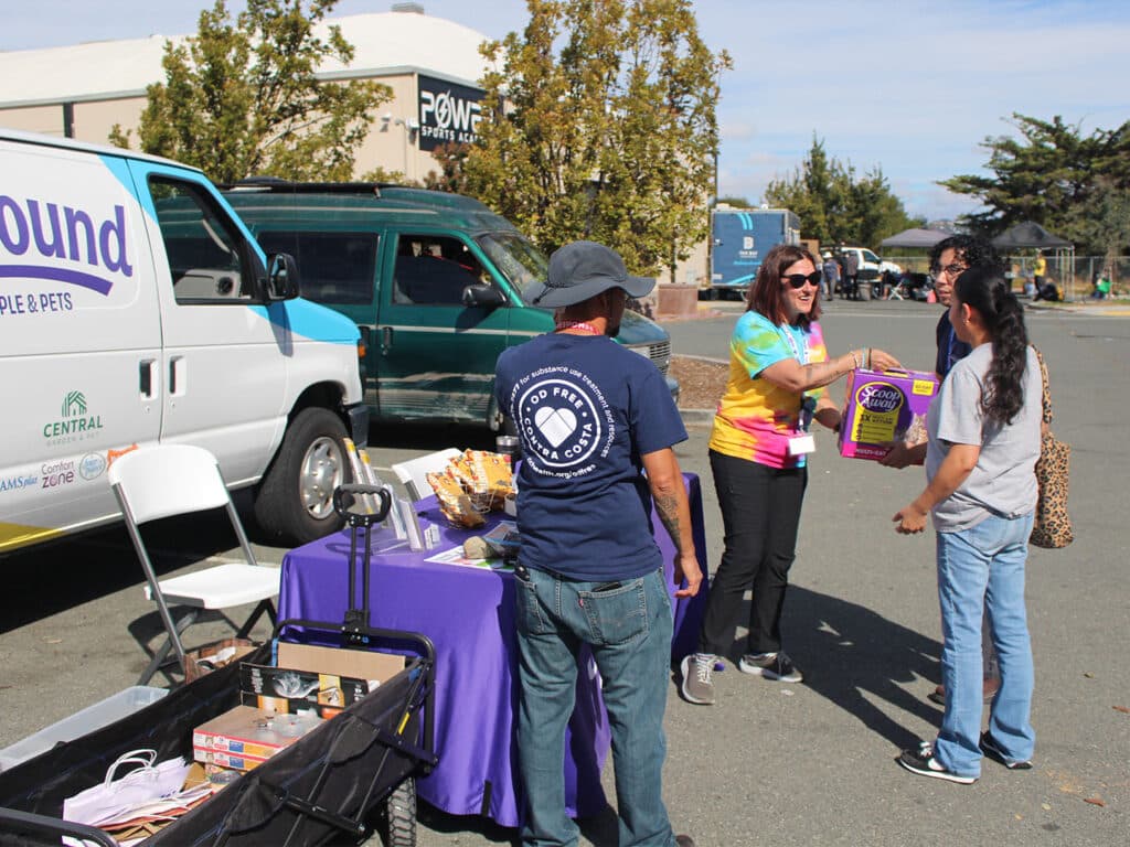 Volunteers and shoppers stand beside tables of pantry supplies in a parking lot, with a white van and parked vehicles in the background.