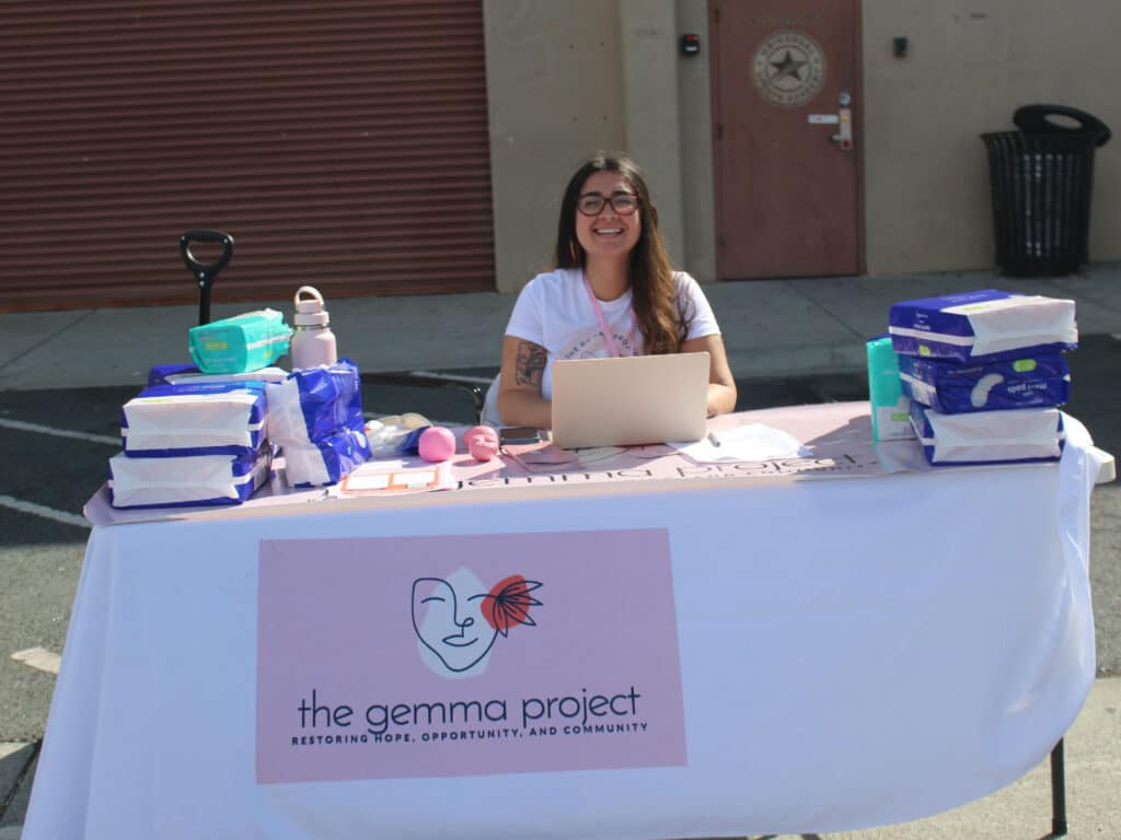 A woman sits behind a table with a laptop and stacks of packaged diapers, with a sign for the Gemma Project on the front and a building entrance in the background.