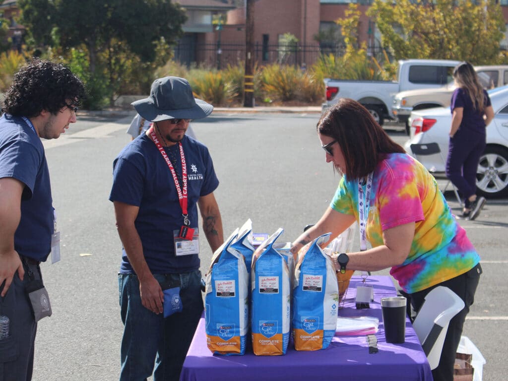 Three volunteers stand at a purple-covered table with bags of food supplies, while a woman in a tie-dye shirt arranges items in a parking lot.