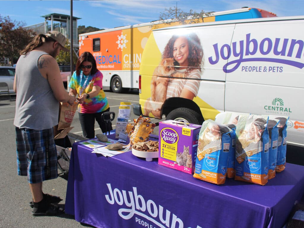 Two people stand beside a purple Joybound People & Pets table with pet food and supplies arranged on top, in front of a branded vehicle parked outdoors.