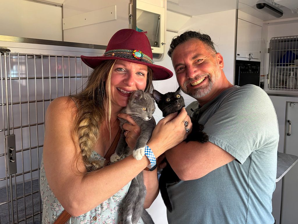 A smiling woman in a red hat holds a gray kitten, while a smiling man holds a black kitten, inside a pet adoption van with metal cages in the background.