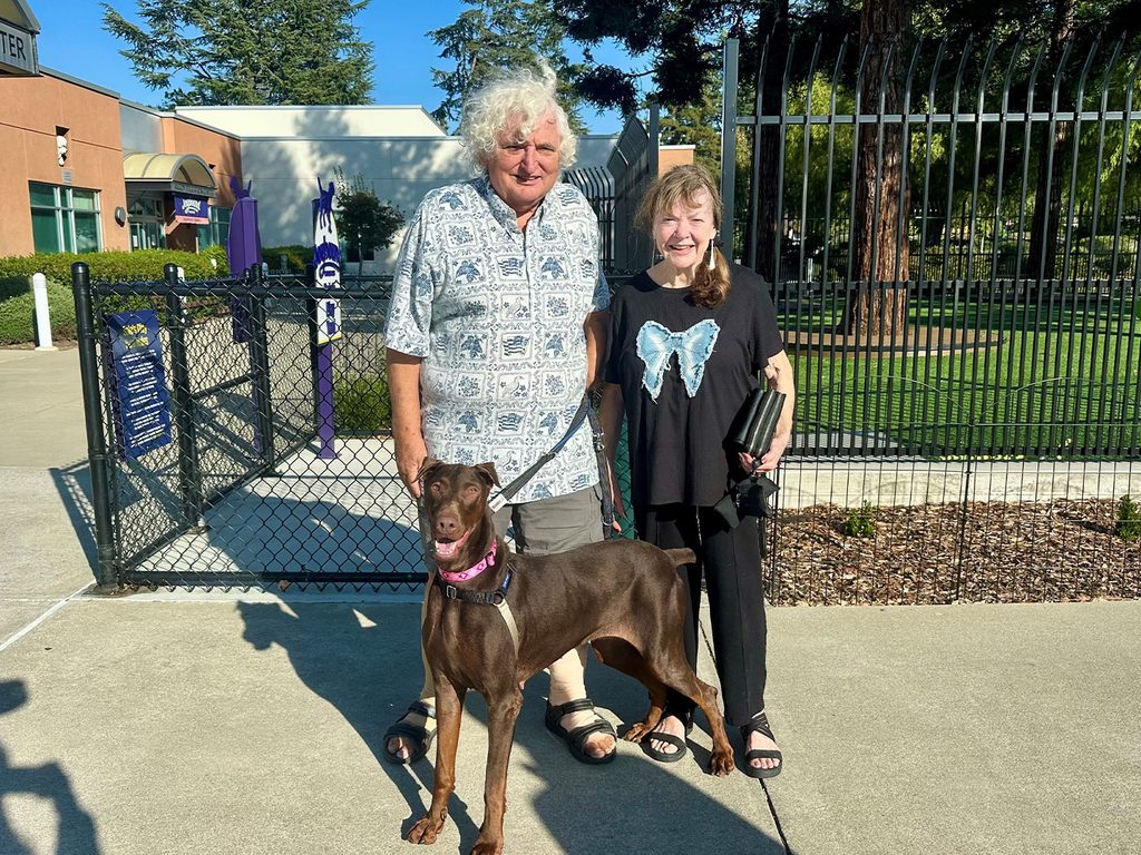 An older man and woman stand with a brown dog wearing a pink collar near a fenced outdoor area, with a building, trees, and clear sky in the background.